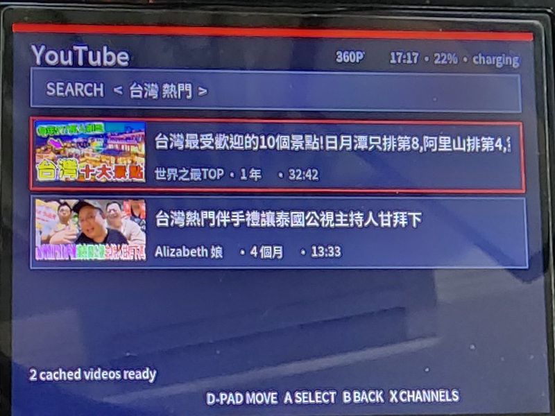 YouTube HOME:搜尋列、兩則影片與縮圖、左下角「2 cached videos ready」、底部按鍵提示 D-PAD MOVE / A SELECT / B BACK / X CHANNELS